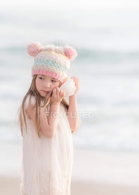 Mädchen steht mit lustigem Hut am Strand und hört einer Muschel zu — Stockfoto