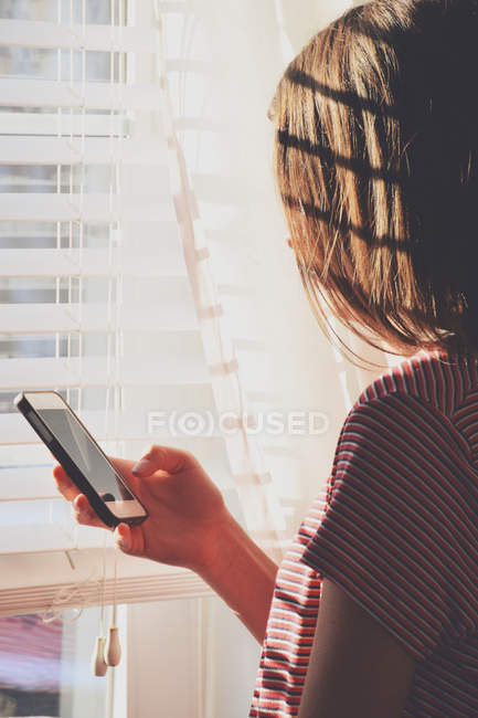Teenage girl standing at window and using mobile phone — Stock Photo