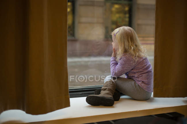 Blond little girl looking out of a window while sitting on window sill — Stock Photo