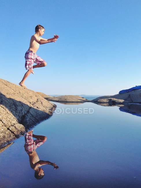Menino pulando de uma rocha no mar com céu azul no fundo — Fotografia de Stock