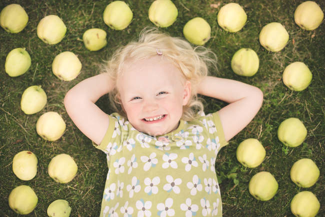 Blondes kleines Mädchen liegt im Gras, umgeben von Äpfeln — Stockfoto