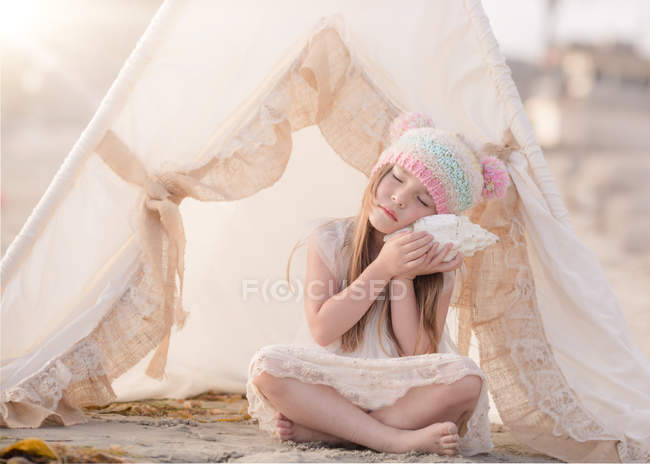 Menina sentada em wigwam na praia e ouvindo uma concha — Fotografia de Stock