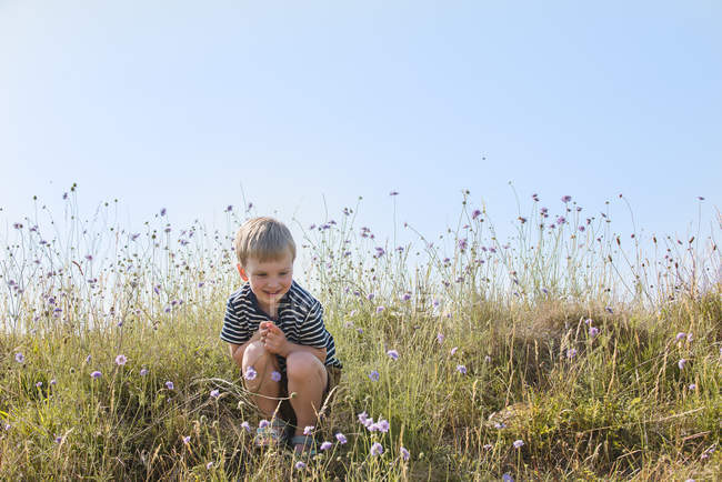 Улыбающийся мальчик сидит в зеленом поле и смотрит на цветы — стоковое фото