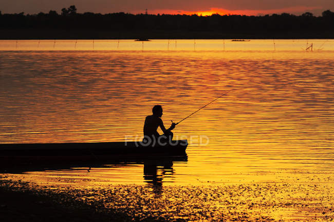 Silueta del hombre sentado en un barco y pescando al atardecer - foto de stock
