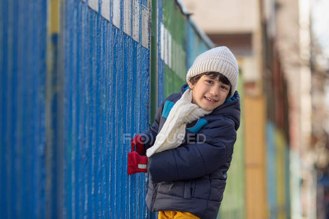 Niño con sombrero sosteniendo una valla de metal y mirando a la cámara - foto de stock