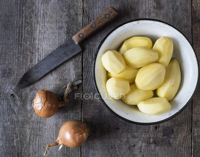 Чистый картофель в миске, лук и нож на деревянном столе — стоковое фото