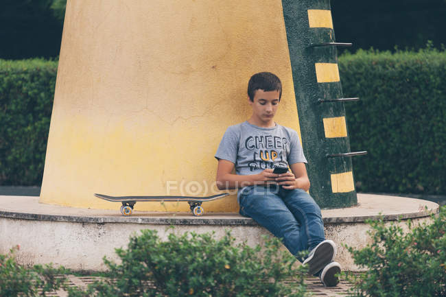 Мальчик сидит со скейтбордом и использует смартфон — стоковое фото