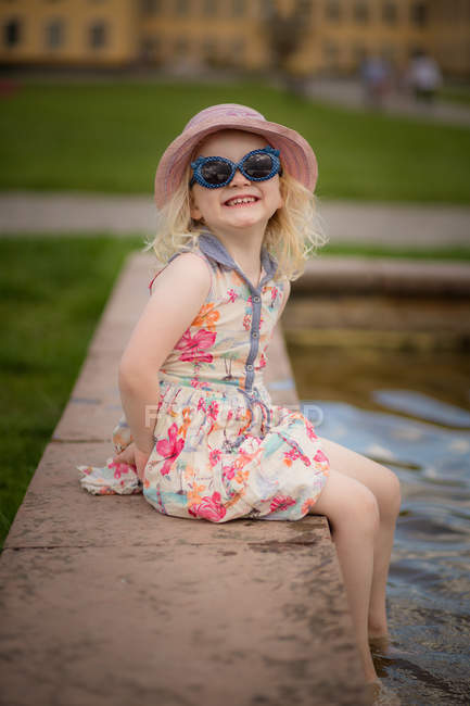 Девушка, сидящая с ногами в воде в узорчатом летнем платье и солнечных очках — стоковое фото