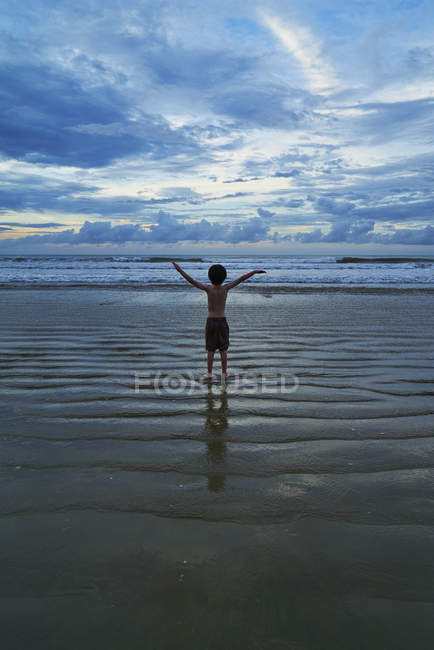 Vue arrière du garçon debout avec les bras en l'air sur la plage — Photo de stock