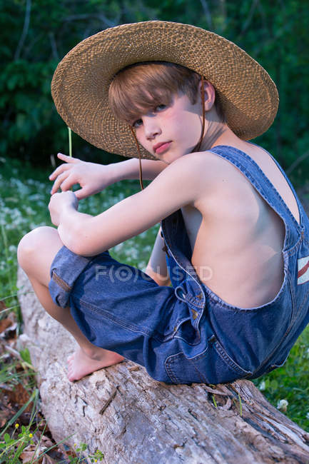 Young boy wearing overalls sitting on tree wearing straw hat — Stock Photo