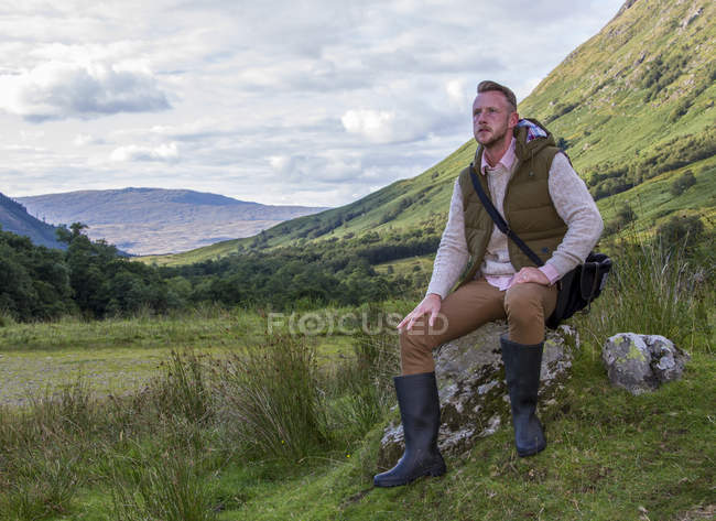 Reino Unido, Escocia, Hombre sentado en la roca en las montañas y mirando hacia otro lado - foto de stock