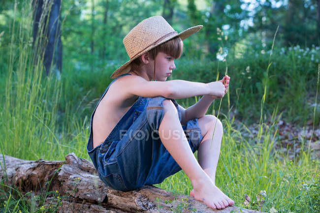Young boy wearing straw hat sitting on log — Stock Photo