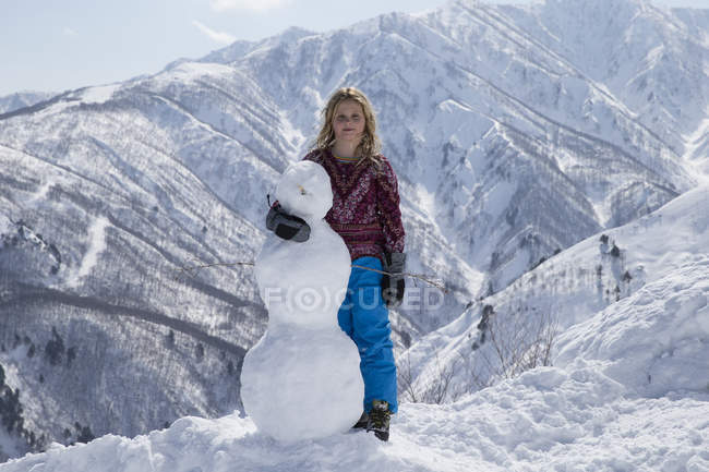 Fille blonde posant avec bonhomme de neige dans les montagnes enneigées — Photo de stock