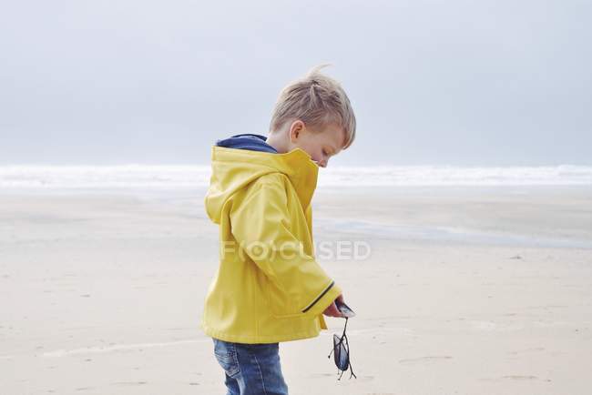 Vue latérale du beau petit garçon en imperméable jaune sur la plage tenant des moules — Photo de stock