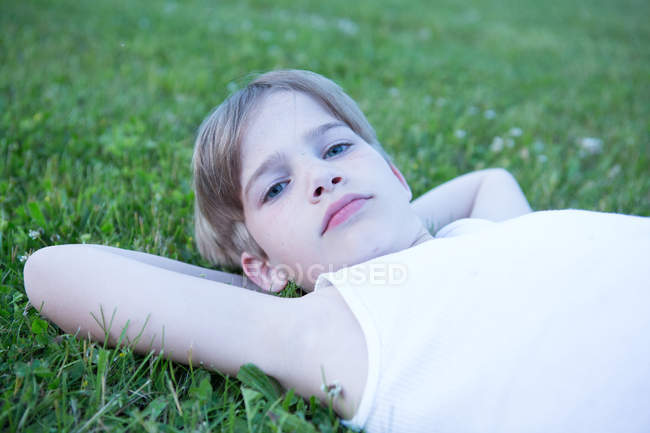 Porträt eines blonden Jungen, der mit den Händen hinter dem Kopf auf dem Gras liegt — Stockfoto
