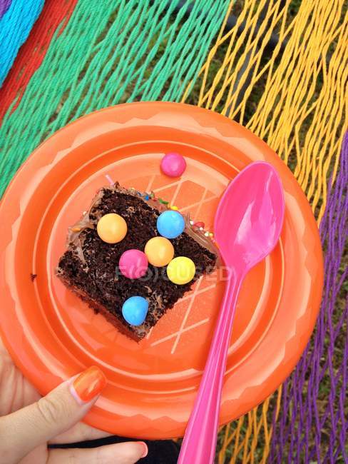 Weibliche Hand hält ein Stück Schokoladenkuchen auf Plastikteller vor buntem Hintergrund — Stockfoto