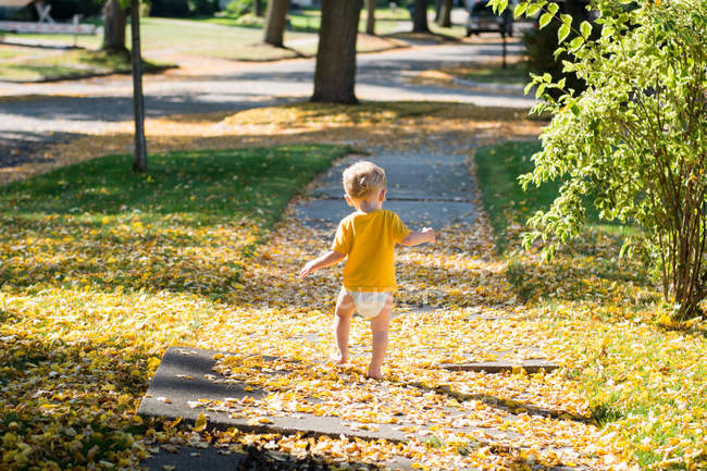 Rückansicht eines Jungen, der durch Herbstblätter auf dem Gehweg läuft — Stockfoto