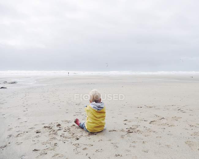 Vue arrière du petit garçon en imperméable jaune assis sur une plage de sable — Photo de stock