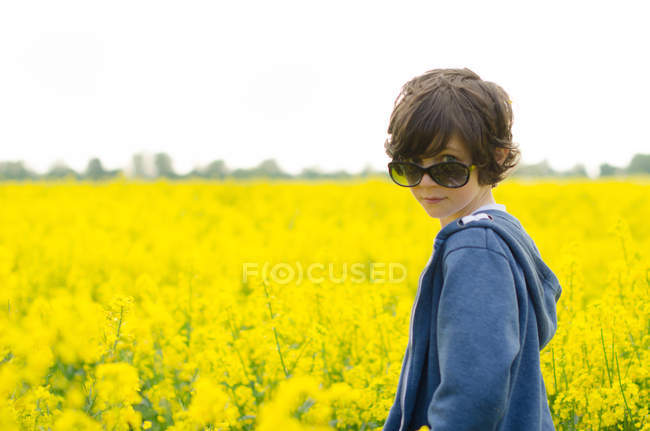 Young boy wearing sunglasses standing against field of yellow flowers — Stock Photo