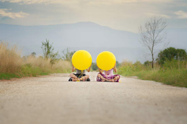 Duas meninas sentadas na estrada rural com balões amarelos — Fotografia de Stock