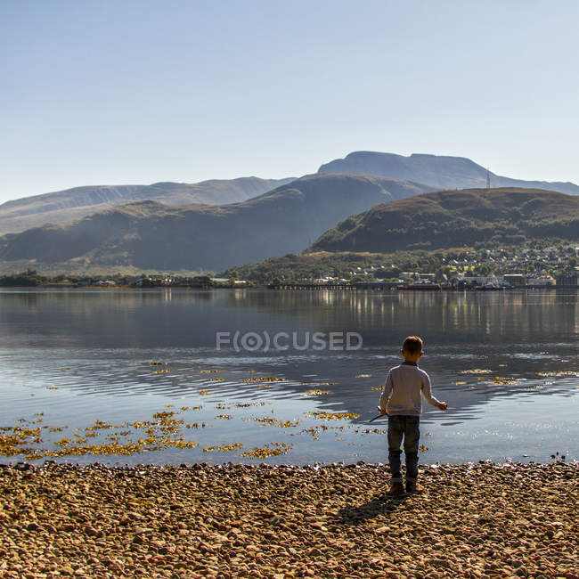 Великобритания, Шотландия, Мальчик бросает камни в воду с горами на заднем плане — стоковое фото