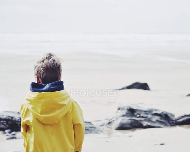 Rear view of little boy in yellow raincoat standing on the beach — Stock Photo