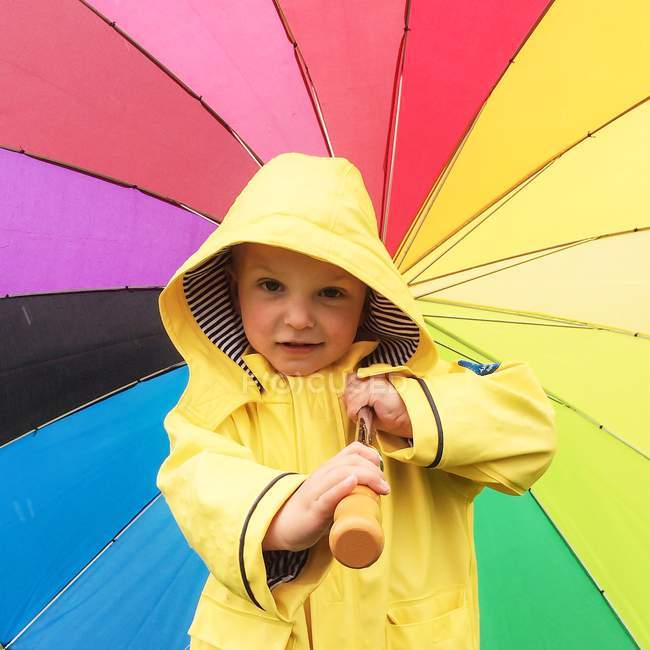 Милий маленький хлопчик в жовтому плащі з барвистою парасолькою дивиться на камеру — стокове фото