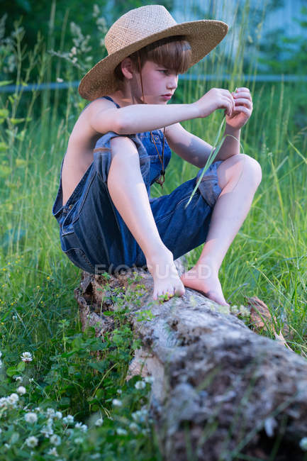 Niño vistiendo un mono azul y sombrero sentado en un árbol caído - foto de stock