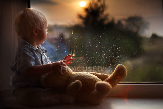 Niño con oso de peluche sentado en el alféizar de la ventana y viendo el atardecer - foto de stock