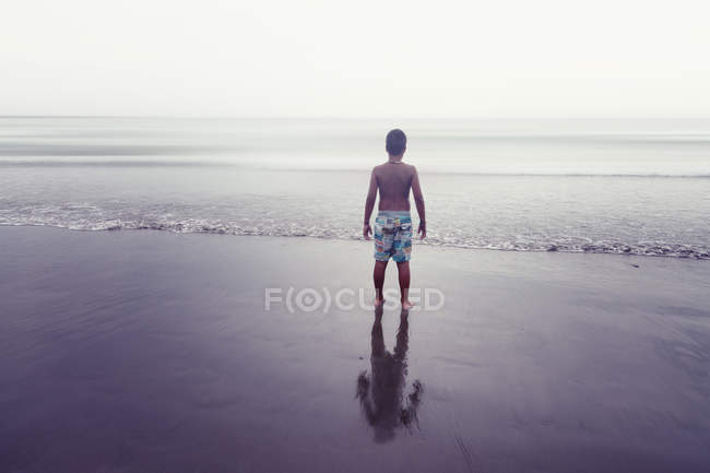 Vista trasera de un chico solitario sin camisa parado en la playa - foto de stock