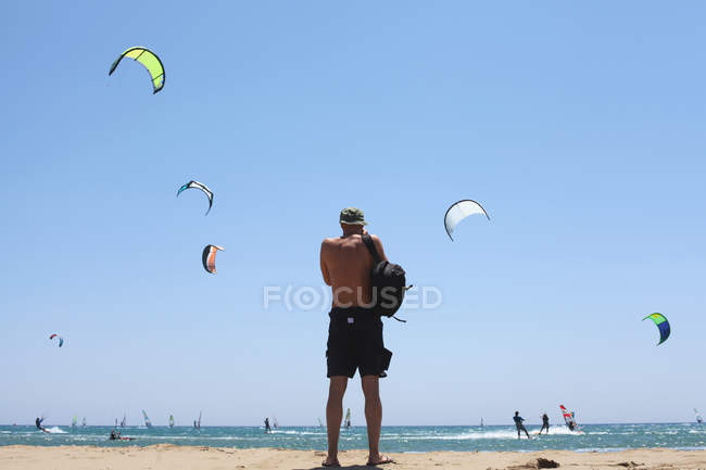 Вид сзади человека, стоящего на пляже и смотрящего на воздушных змеев — стоковое фото