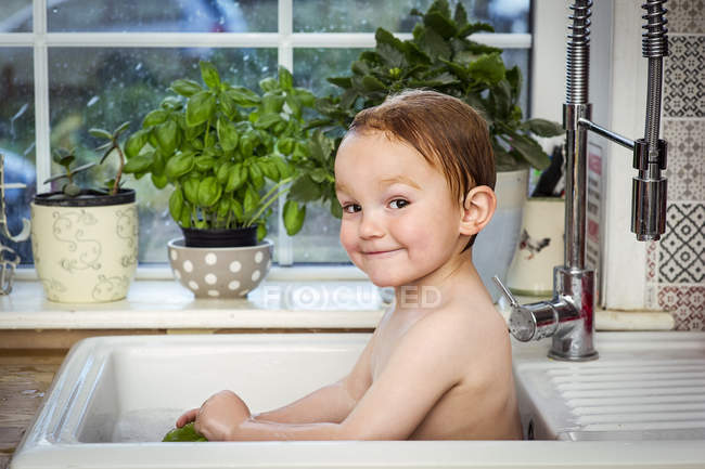 Carino bambino che fa il bagno nel lavandino della cucina e guarda la fotocamera — Foto stock
