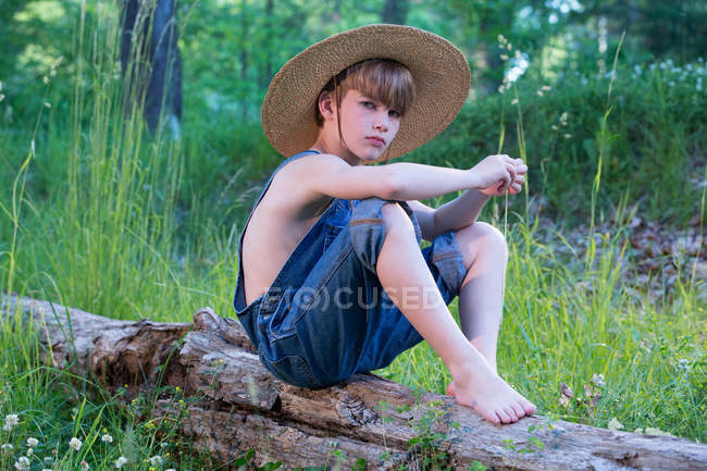 Young boy wearing overalls sitting on tree wearing straw hat — vertical ...