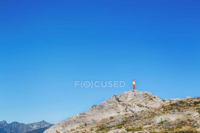 Ragazzo in piedi sulla cima di una collina rocciosa con cielo blu su sfondo — Foto stock