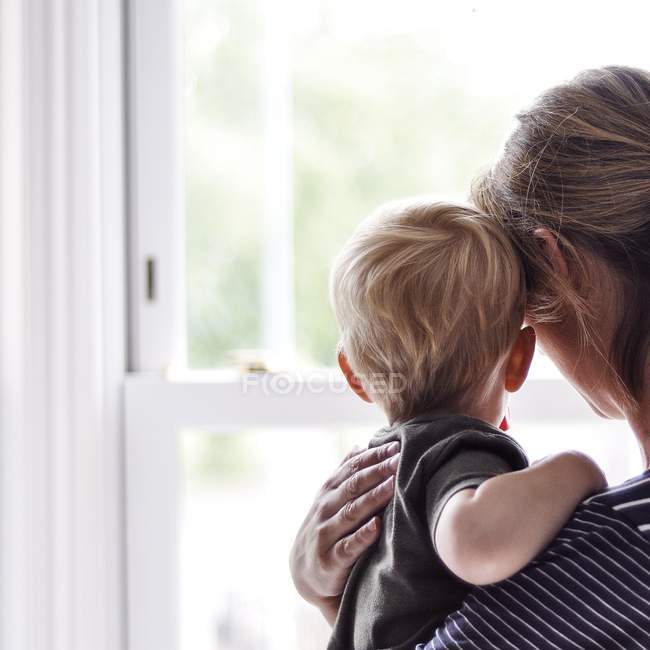 Visão traseira da mãe e do filho pequeno olhando pela janela — Fotografia de Stock