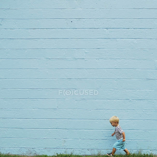 Garçon marchant sur la pelouse devant le mur de briques bleues — Photo de stock