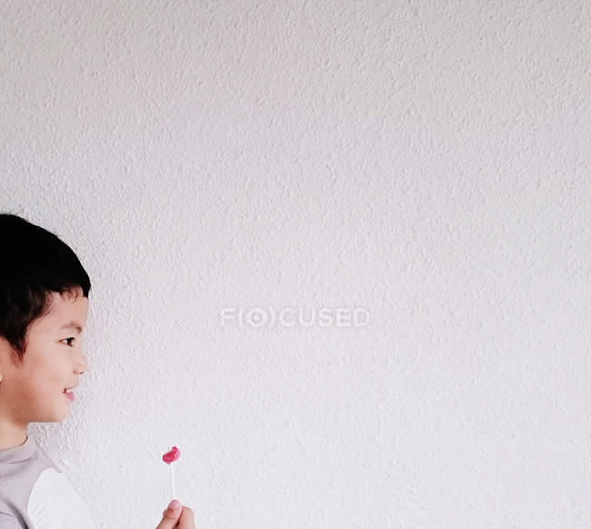 Sonriente chico sosteniendo mitad comido piruleta en frente de blanco pared - foto de stock
