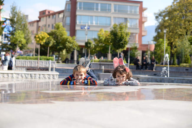 Cute girl and boy lying on ground at city square and looking at camera — Stock Photo