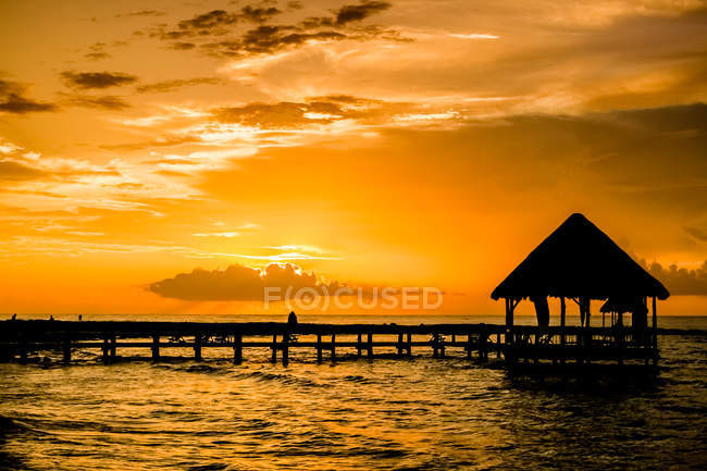 Hermoso cielo naranja atardecer, agua de mar con muelle y pequeña cabaña - foto de stock