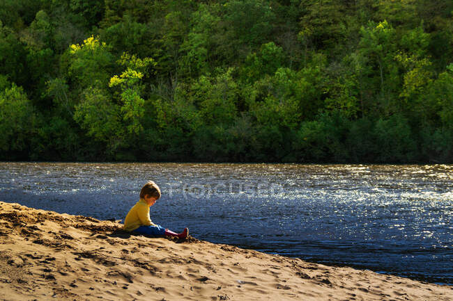 Niño solitario sentado en la orilla del río en el día soleado - foto de stock