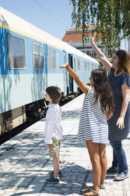 Madre y dos niños de pie en la estación de tren saludando - foto de stock
