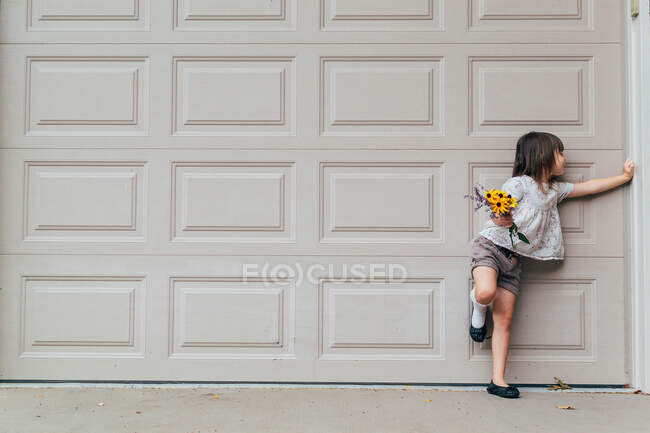 Fille debout devant la porte de garage tenant des fleurs — Photo de stock