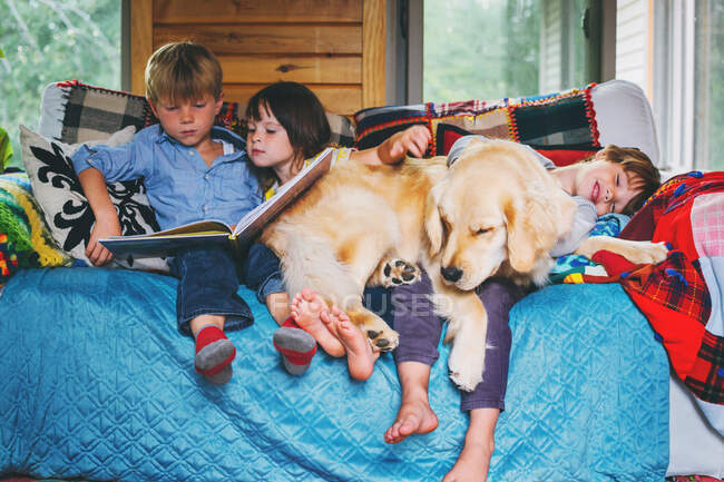 Trois enfants assis sur le canapé lisant avec le chien — Photo de stock