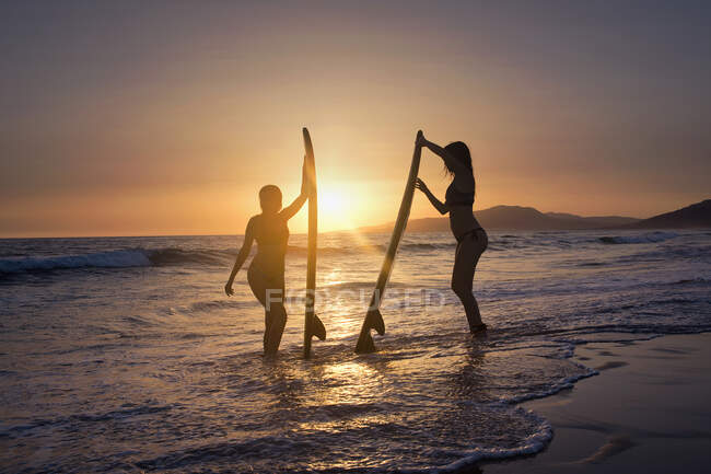 Silueta de Dos mujeres de pie en el océano con tablas de surf, Playa de Los Lances, Tarifa, Cádiz, Andalucía, España - foto de stock