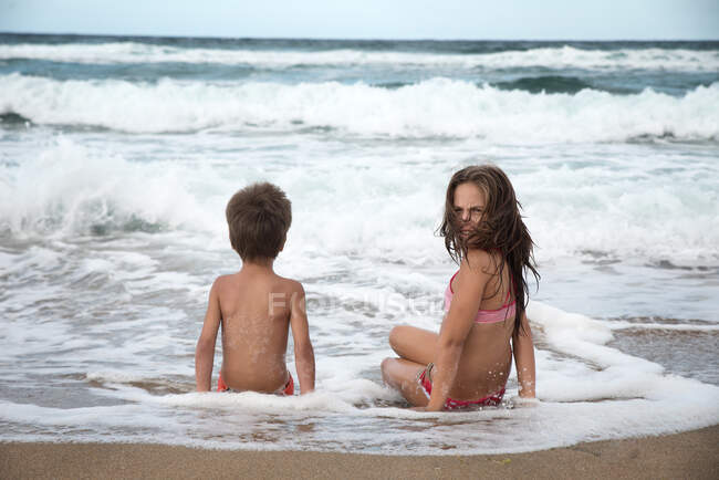 Girl and boy sitting on beach in water's edge — Stock Photo