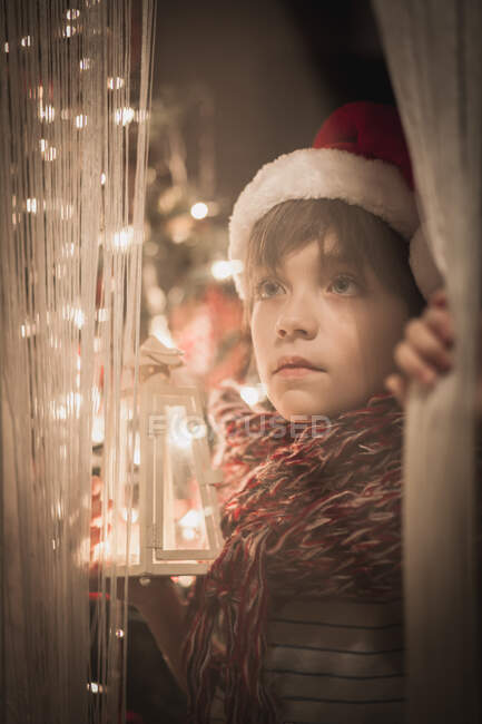 Мальчик у окна в рождественской шляпе Санты с фонариком — стоковое фото