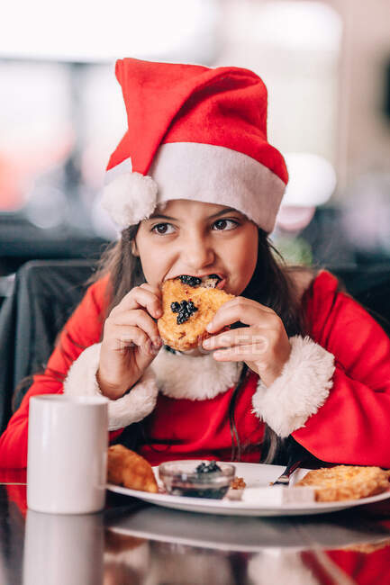 Menina em uma roupa de Natal Papai Noel e chapéu comendo café da manhã — Fotografia de Stock