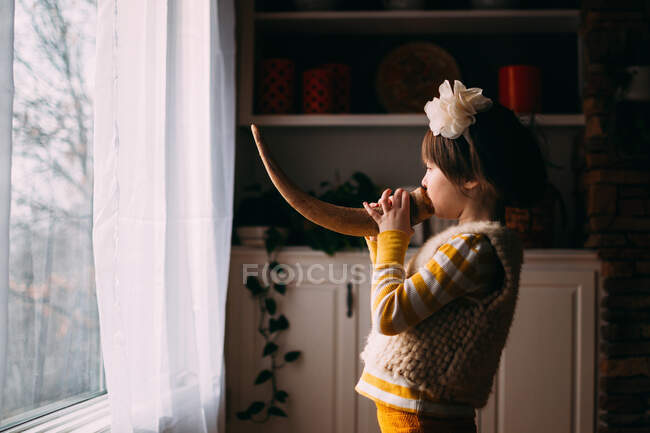 Chica de pie frente a la ventana tocando un cuerno - foto de stock