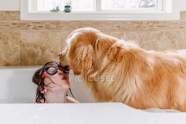 Girl wearing swimming goggles in the bath with her golden retriever dog — Stock Photo