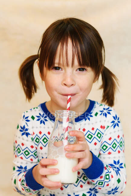 Retrato de una niña en pijama bebiendo una pequeña botella de leche a través de una paja - foto de stock